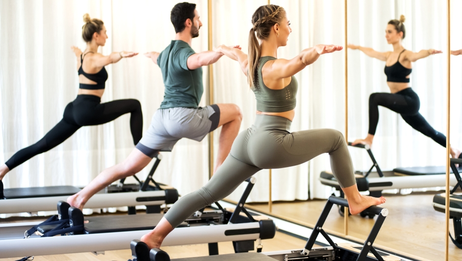 Class in a gym doing pilates standing lunges on reformer beds to stretch and tone the muscles reflected in a wall mirror