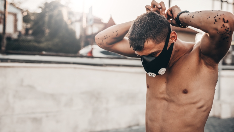 Caucasian muscular male wearing black training mask to increase load on breathing muscles during morning cardio workout routine outdoors. Copy space. Shirtless sportsman doing workout exercise outside