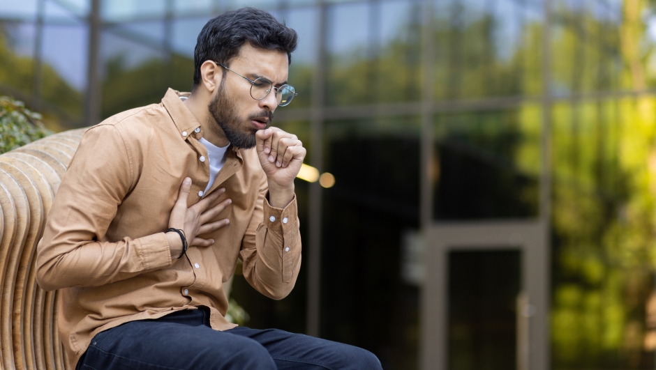 Young man wearing glasses and casual clothing appears to be coughing, indicating possible respiratory issues, allergy, or health problem. Image captures moment of discomfort in urban outdoor setting
