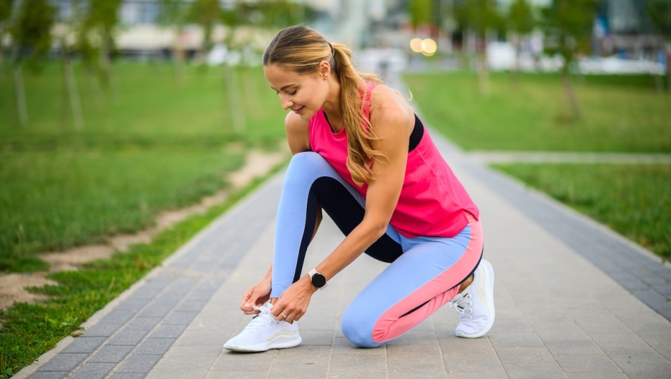 Young athletic woman kneeling on the grass, tying shoelaces on her running shoes, getting ready for a jog in the city park, focused on her fitness and training routine