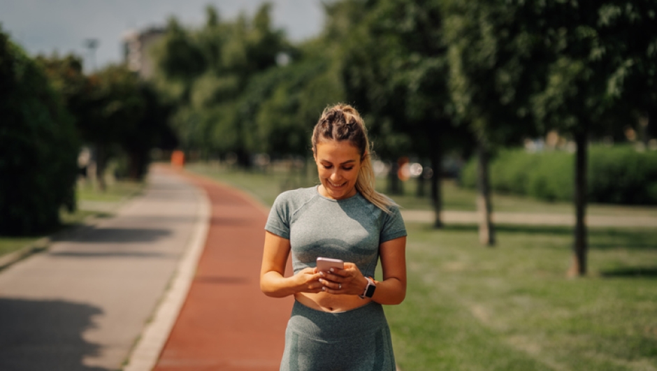 Fit woman engaging with a mobile phone app to track workout performance while running along a scenic path in a sunny park