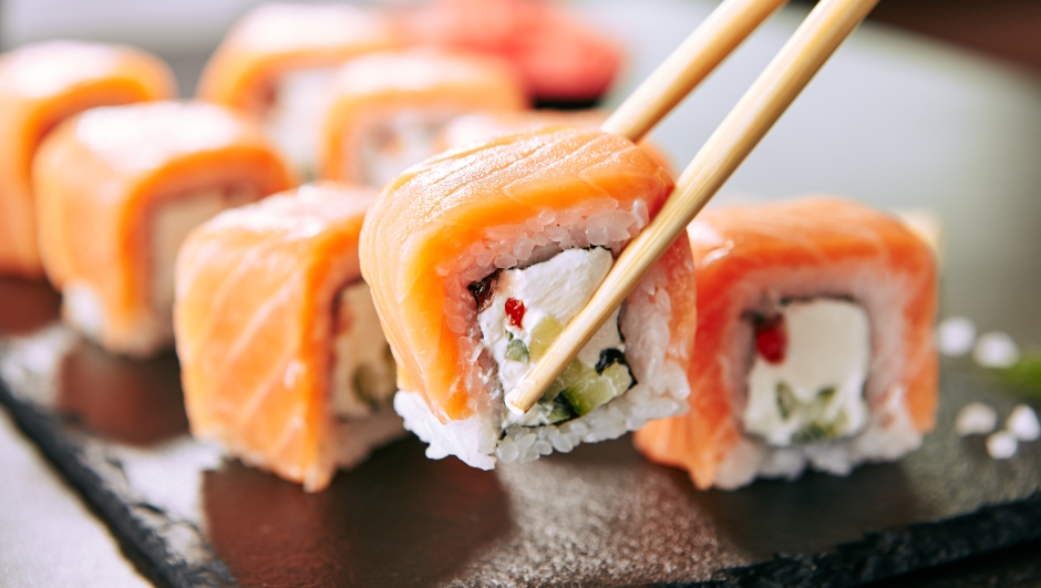 Chopstick holding Sushi Rolls Set with Salmon and Cream Cheese and Cuccumber on Black Slate Plate Close Up. Uramaki, Nori Maki or Futomaki Sushi with Trout Fillets, Soy Sauce and Wasabi