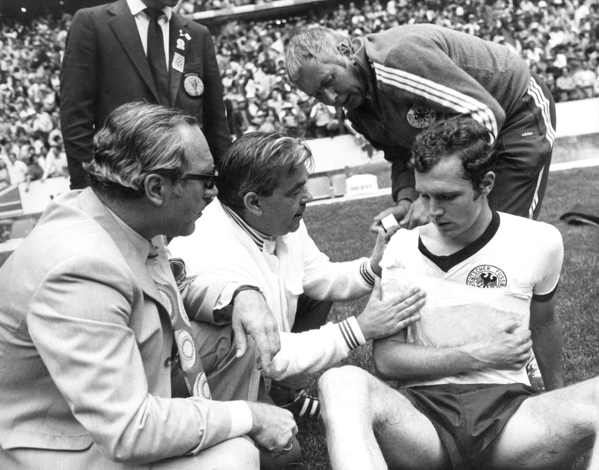 West German defender Franz Beckenbauer (R) gets his right shoulder bandaged by a masseur as the team's doctor Professor Schoberth (L) looks on during the World Cup semifinal soccer match against Italy 17 June 1982 at Azteca stadium in Mexico City. At the end of regulation time, the two teams were tied at 1 and Beckenbauer went on to play during the thirty-minute extra time period but could not prevent his team from losing 4-3 on a late goal by midfielder Gianni Rivera.    AFP PHOTO/EPA/DPA (Photo by DPA / dpa Picture-Alliance via AFP)
