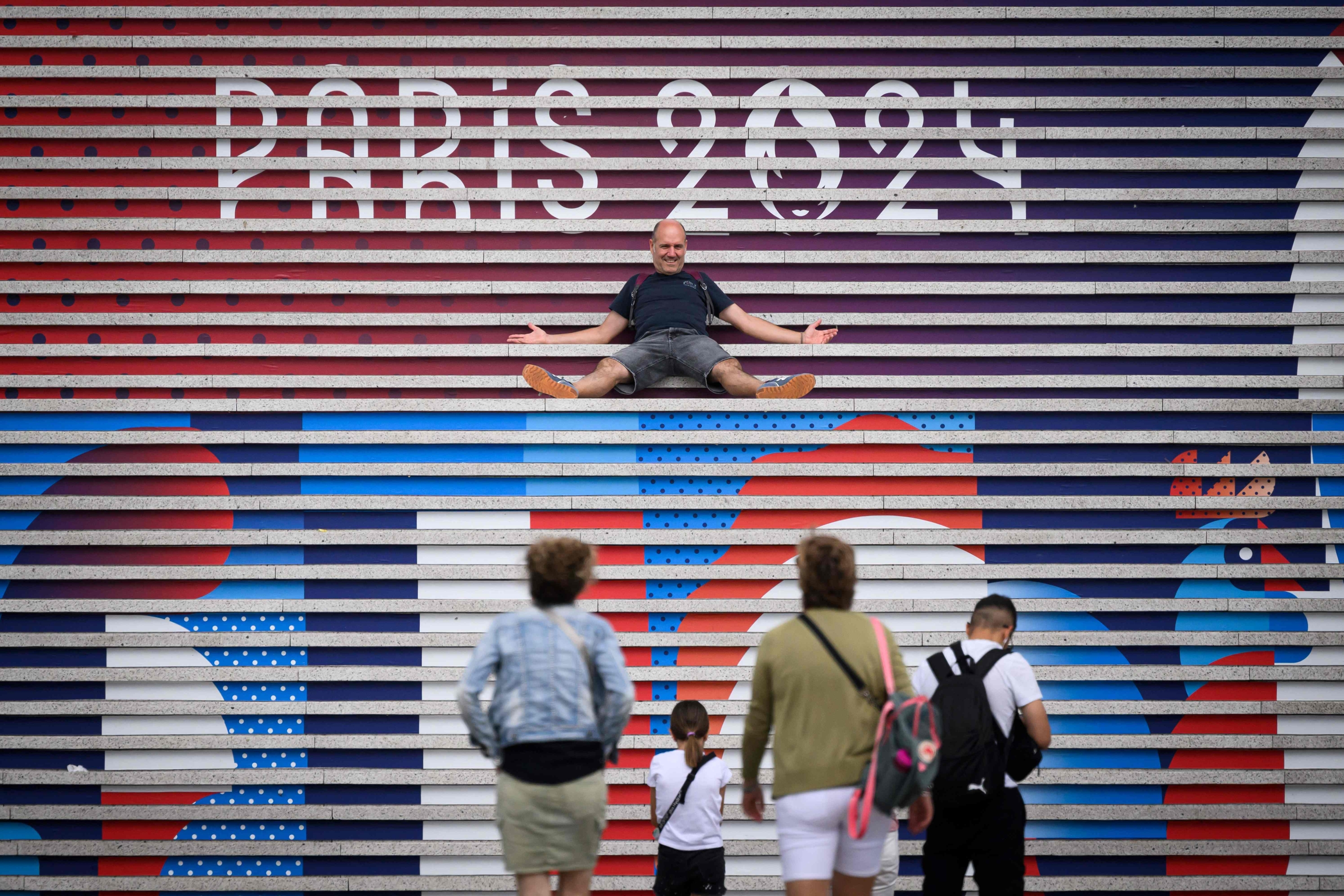 People pose on stairs near the logo of Paris 2024 Olympics Olympic games a few days before the opening ceremony, at La Defense buisiness district, West of Paris, on July 22, 2024. (Photo by Loic VENANCE / AFP)