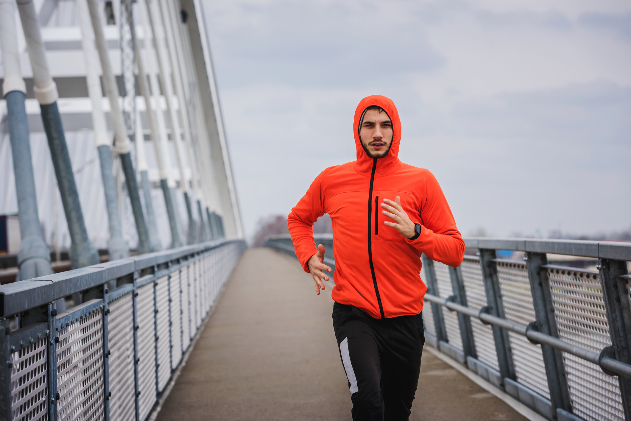 Handsome young athlete running fast along big modern bridge in orange windbreaker jacket. Exercising, Jogging, Sport, Winter. Male athlete running.