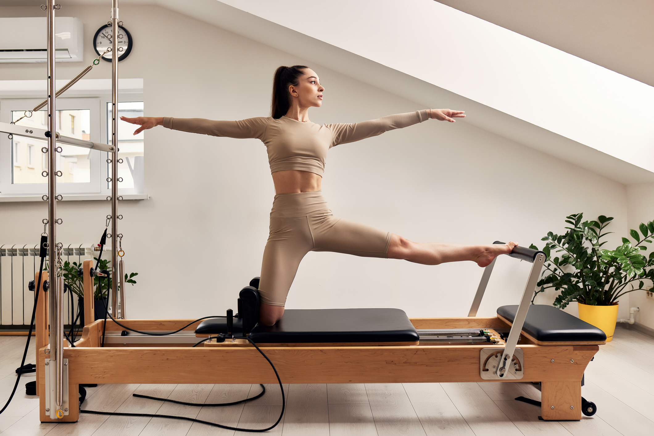 Young girl is doing Pilates on a reformer bed in a bright studio. Slender brunette in a beige bodysuit does exercise to train balance and stretch the muscles of her legs. Healthy lifestyle concept