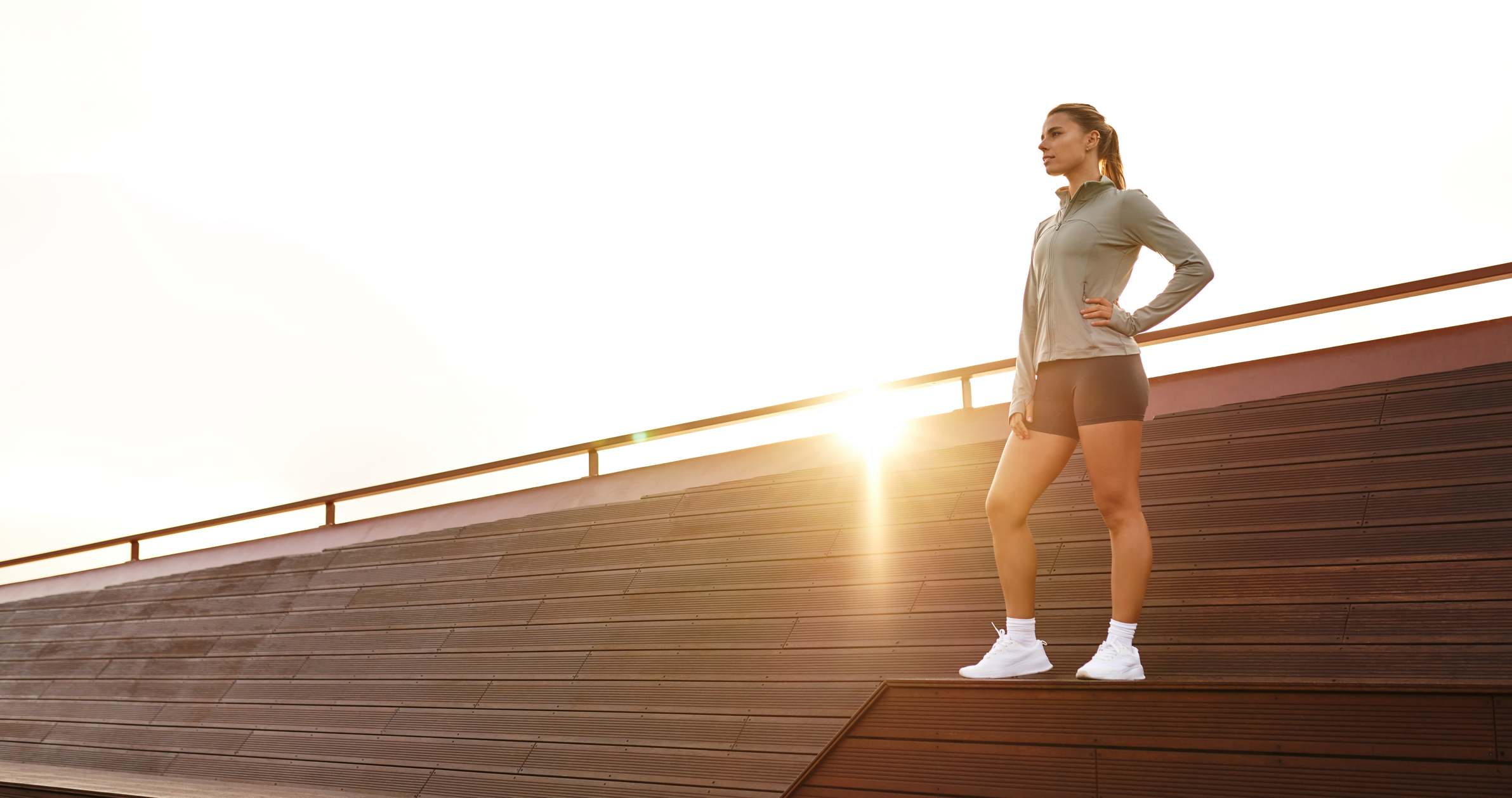 A confident, athletic woman stands tall in an assertive pose on a rooftop at sunset, showcasing her strength and determination in a serene urban setting filled with beauty and calmness